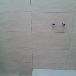 Ściana łazienki wyłożona beżowymi płytkami ceramicznymi z widocznymi otworami na instalację wodną i fragmentem wanny u dołu.