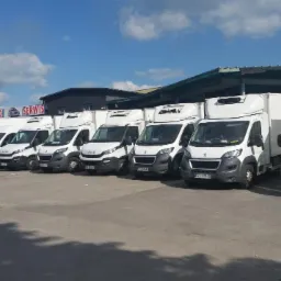 Flota białych samochodów dostawczych różnych marek (Iveco, Peugeot) z nadbudówkami chłodniczymi, zaparkowana na placu pod zadaszeniem, w tle budynek serwisu samochodowego i błękitne niebo z chmurami.