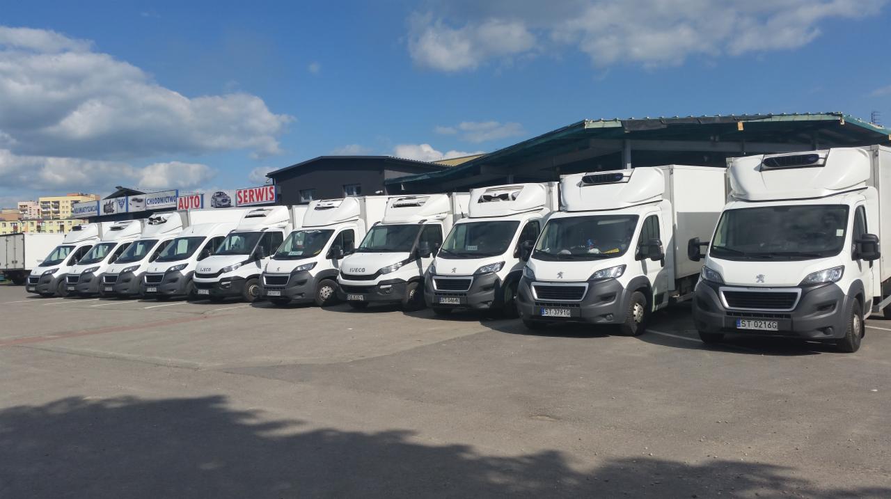 Flota białych samochodów dostawczych różnych marek (Iveco, Peugeot) z nadbudówkami chłodniczymi, zaparkowana na placu pod zadaszeniem, w tle budynek serwisu samochodowego i błękitne niebo z chmurami.