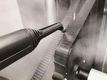 Dysza parowa czyszcząca taśmę transportową w zakładzie przemysłowym, widoczna para i elementy mechaniczne.