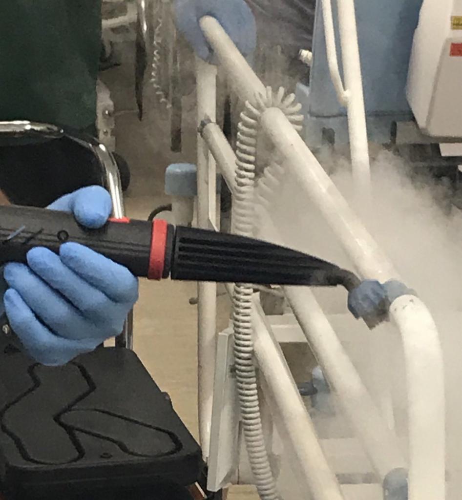 Czyszczenie parowe metalowej konstrukcji łóżka szpitalnego za pomocą parownicy trzymanej w dłoniach w niebieskich rękawicach ochronnych.