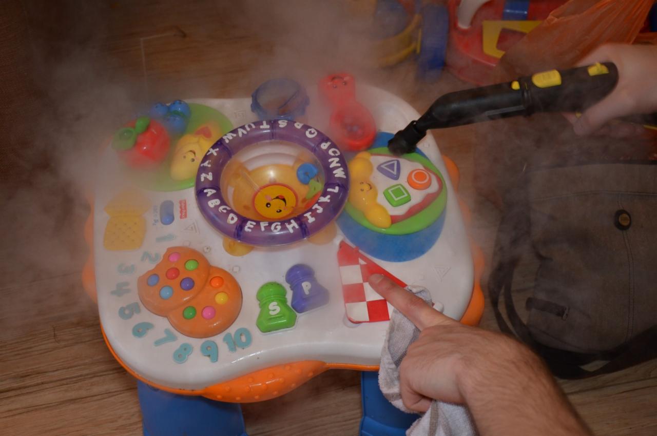 Czyszczenie parowe interaktywnego stolika edukacyjnego dla dzieci, para wydobywa się z dyszy parownicy, dłoń przeciera powierzchnię szmatką, widoczny fragment podłogi i torby.