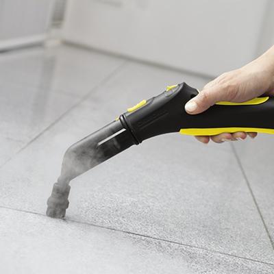 Czyszczenie fug na szarych płytkach podłogowych za pomocą czarno-żółtego parowego urządzenia do czyszczenia, z dyszy wydobywa się para.