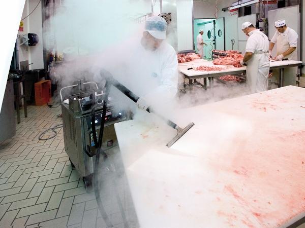 Pracownik w kombinezonie ochronnym czyści parą przemysłowy stół z resztek mięsa w chłodni, widoczny parowy odkurzacz przemysłowy i płytki na podłodze.