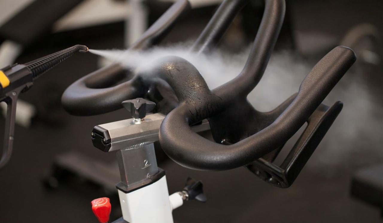 Czyszczenie parowe kierownicy roweru stacjonarnego w siłowni za pomocą dyszy parowej, widoczne krople wody i para.