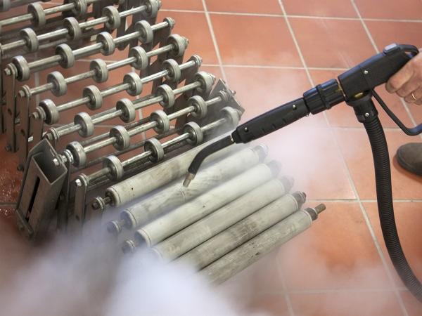 Czyszczenie parowe stalowych rolek transportowych za pomocą pistoletu ciśnieniowego na tle pomarańczowych płytek podłogowych.