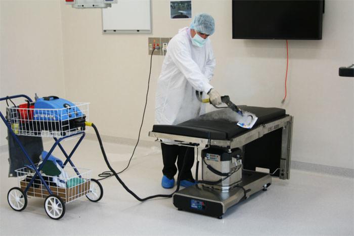 Pracownik w kombinezonie ochronnym czyści parą medyczny stół operacyjny w sterylnym pomieszczeniu szpitalnym, w tle wózek z urządzeniem czyszczącym.