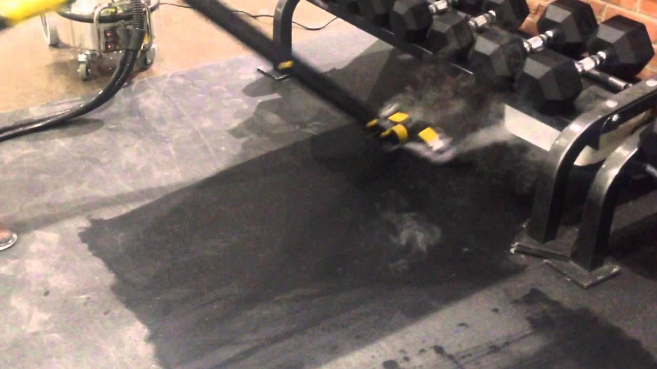 Czyszczenie parowe gumowej podłogi w siłowni, widoczny sprzęt do ćwiczeń i para wydobywająca się spod dyszy czyszczącej.