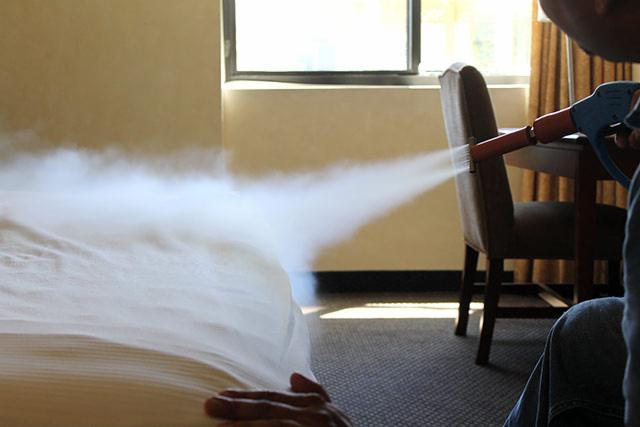 Dezynfekcja parowa hotelowego łóżka za pomocą specjalistycznego urządzenia, widoczna para, krzesło i biurko w tle, osoba w jeansach obsługująca urządzenie.