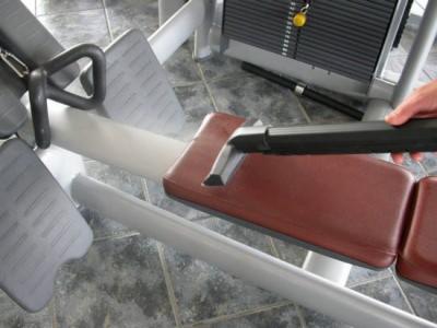 Czyszczenie tapicerowanego siedziska maszyny do ćwiczeń w siłowni za pomocą odkurzacza z wąską ssawką.