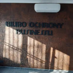 Ściana w biurze o fakturze w odcieniach brązu z napisem 'BIURO OCHRONY BUSINESSU' wykonanym z czarnych liter, oświetlona promieniami słońca wpadającymi przez okno, z zamontowanym klimatyzatorem.