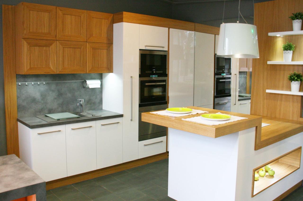 Nowoczesna kuchnia z białymi frontami i drewnianymi akcentami, wyspa z blatem drewnianym, wisząca lampa i półki z roślinami doniczkowymi.