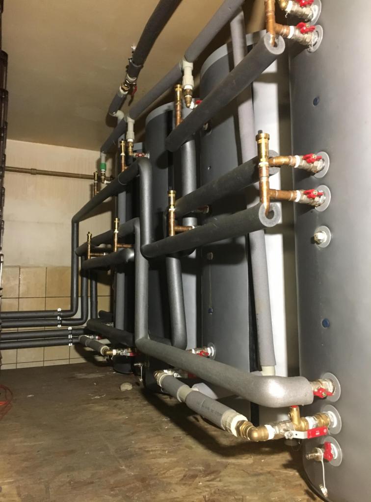 Szeregowo połączone, zaizolowane zbiorniki akumulacyjne ciepła z licznymi przyłączami i zaworami odcinającymi w pomieszczeniu technicznym.