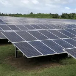 Rozległa farma paneli słonecznych na zielonym trawiastym terenie, z budynkiem w oddali pod pochmurnym niebem.
