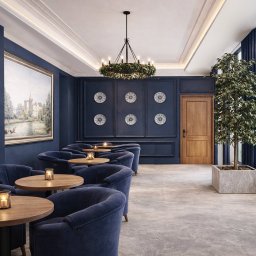 ROLI.ART AGNIESZKA ROLIRAD - Eleganckie wnętrze kawiarni w odcieniach granatu z drewnianymi stolikami, miękkimi fotelami i dekoracyjnym oświetleniem. Na ścianie obraz i ozdobne talerze.