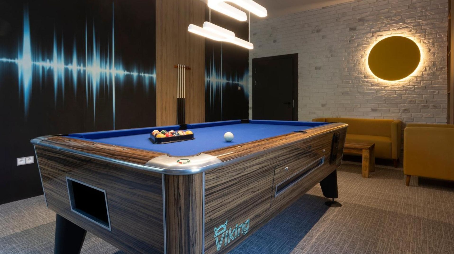 Stylowy pokój gier z drewnianym stołem bilardowym, niebieskim suknem, nowoczesnym oświetleniem i ścianą z motywem dźwiękowym. Hotelowy design.
