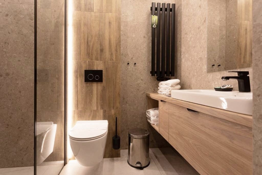 Nowoczesna łazienka z drewnopodobnymi płytkami, podwieszaną toaletą z czarnym przyciskiem spłukiwania, czarnym grzejnikiem drabinkowym z etykietą, umywalką nablatową i drewnianą szafką z zapasem...