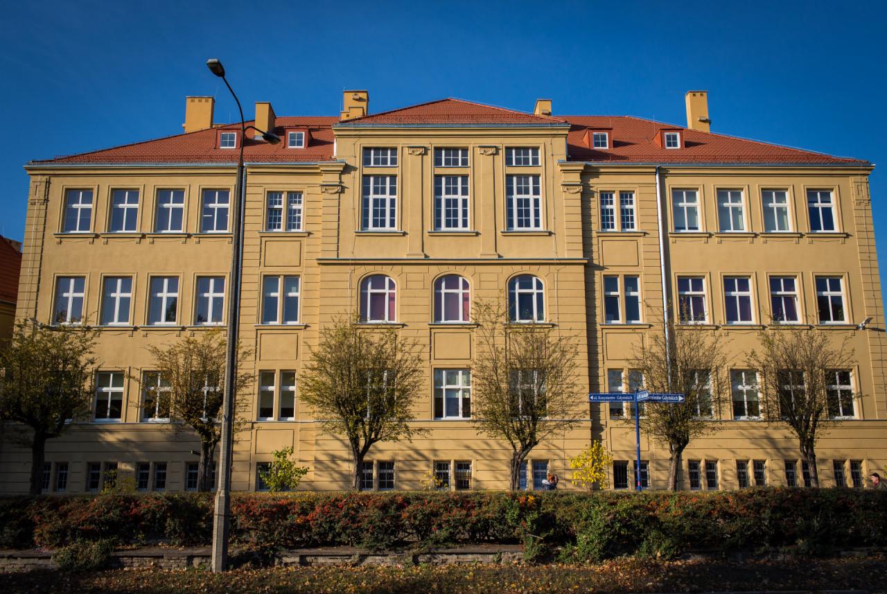 Trzykondygnacyjny budynek o żółtej elewacji z czerwonym dachem, widoczny z poziomu ulicy, z drzewami i krzewami przed budynkiem.