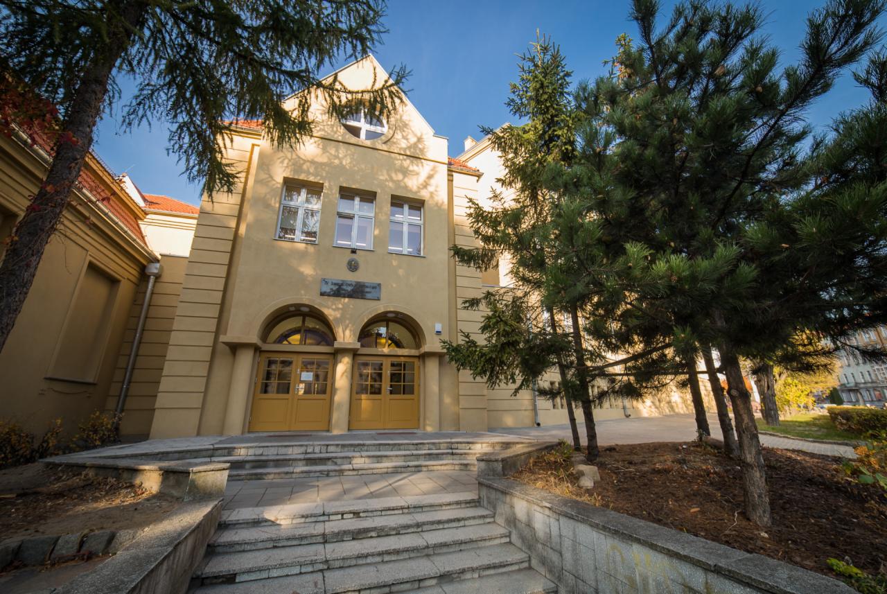 Odnowiony budynek z żółtą elewacją, widoczne schody wejściowe i drzewa iglaste na pierwszym planie.