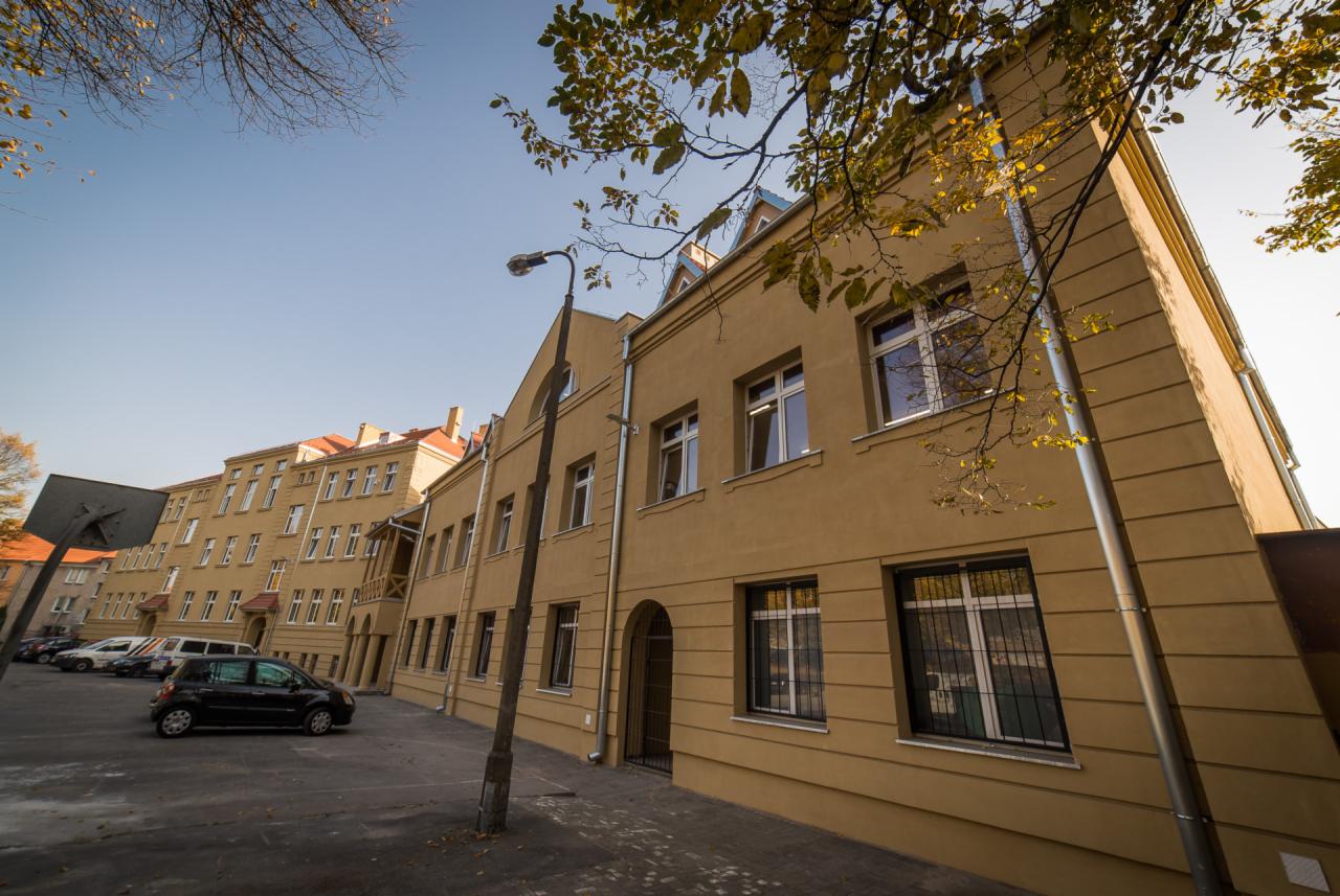 Budynek wielorodzinny z oknami z kratami, otynkowany na kolor piaskowy, widoczny z perspektywy ulicy z zaparkowanymi samochodami i drzewami z jesiennymi liśćmi.