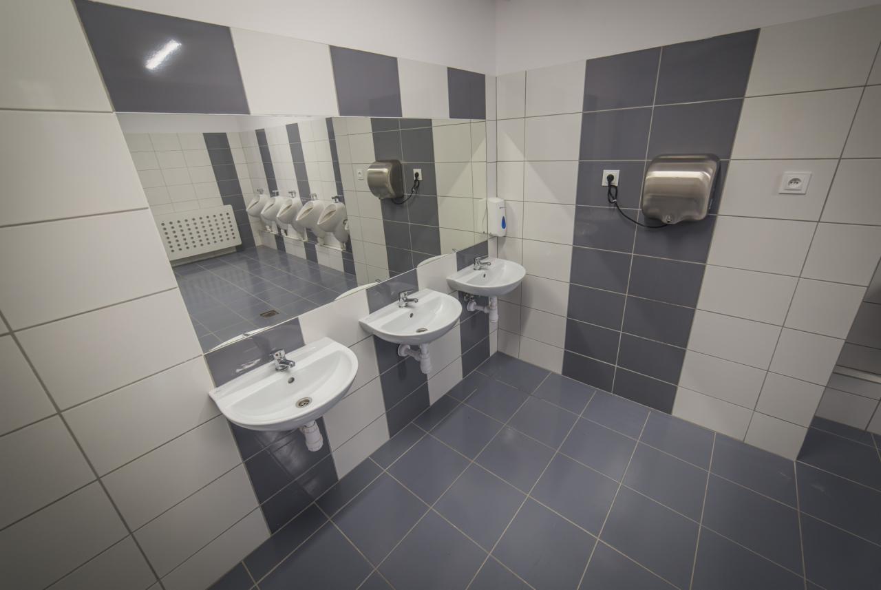 Wykończona łazienka publiczna z umywalkami, suszarką do rąk i pisuarami widocznymi w lustrze, utrzymana w szaro-białej kolorystyce płytek.