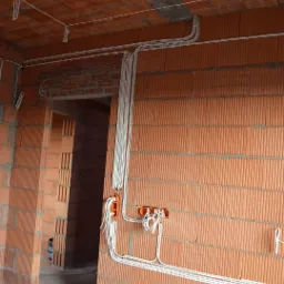 Świeżo położone przewody elektryczne w nowym budynku z cegły, prowadzone wzdłuż ścian i sufitu, widoczne pomarańczowe puszki instalacyjne z wystającymi kablami.