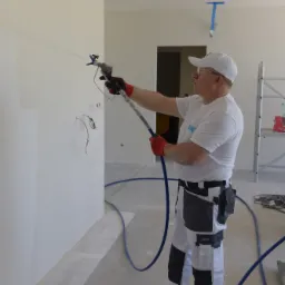 Pracownik w okularach ochronnych i roboczym stroju nanosi natryskowo białą substancję na ścianę w pomieszczeniu w stanie surowym, widoczne wystające kable elektryczne i niebieski wąż doprowadzający...