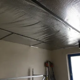 Montaż izolacji termicznej na suficie z widocznymi profilami mocującymi, drabina i rusztowanie w tle.