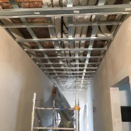 Montaż metalowego stelaża sufitu podwieszanego z widocznymi rurami wentylacyjnymi nad nim, widok z dołu ze stojącego rusztowania.