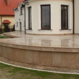 Kamienny taras z beżowego granitu, z niskim murkiem, przylegający do budynku w Lubartowie, z widocznym fragmentem ogrodu i drugiego domu w tle.