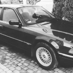 Czarno-białe zdjęcie klasycznego BMW serii 7 E32 zaparkowanego na brukowanej nawierzchni z widocznymi aluminiowymi felgami i odblaskami na karoserii.