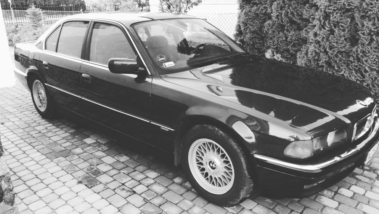 Czarno-białe zdjęcie klasycznego BMW serii 7 E32 zaparkowanego na brukowanej nawierzchni z widocznymi aluminiowymi felgami i odblaskami na karoserii.