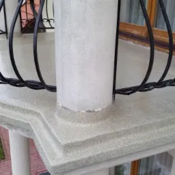 Odnowiony balkon z balustradą z kutego żelaza i jasną, chropowatą powierzchnią posadzki, widok z góry na zielony trawnik i fragment czerwonej cegły.
