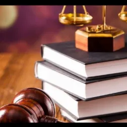 Drewniany stół z trzema książkami w twardej oprawie, na których leżą złote wagi sprawiedliwości i drewniany młotek sędziowski
