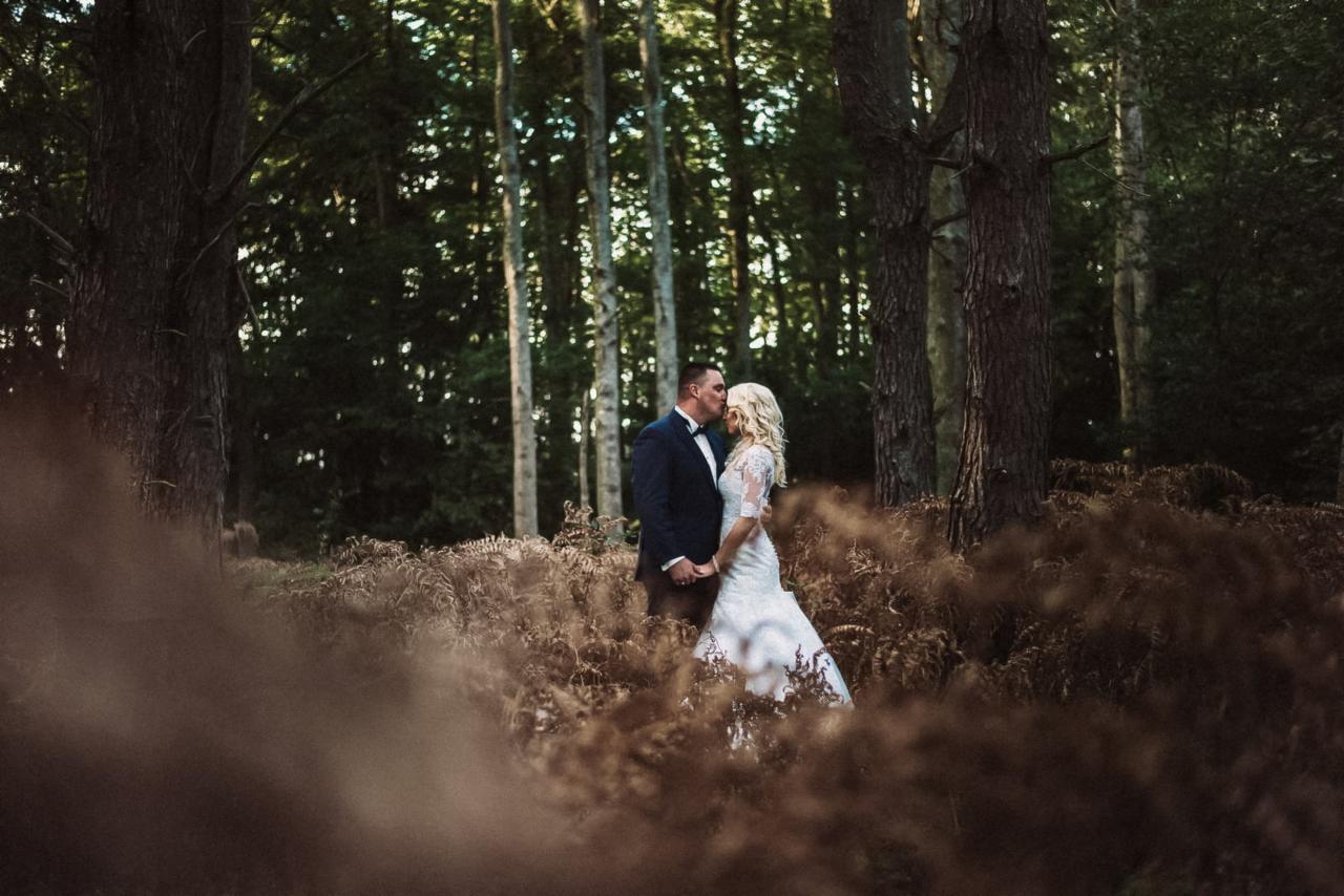 Para młoda w lesie, panna młoda w białej sukni z koronkowymi rękawami, pan młody w granatowym garniturze z muszką, całujący się na tle drzew i paproci.