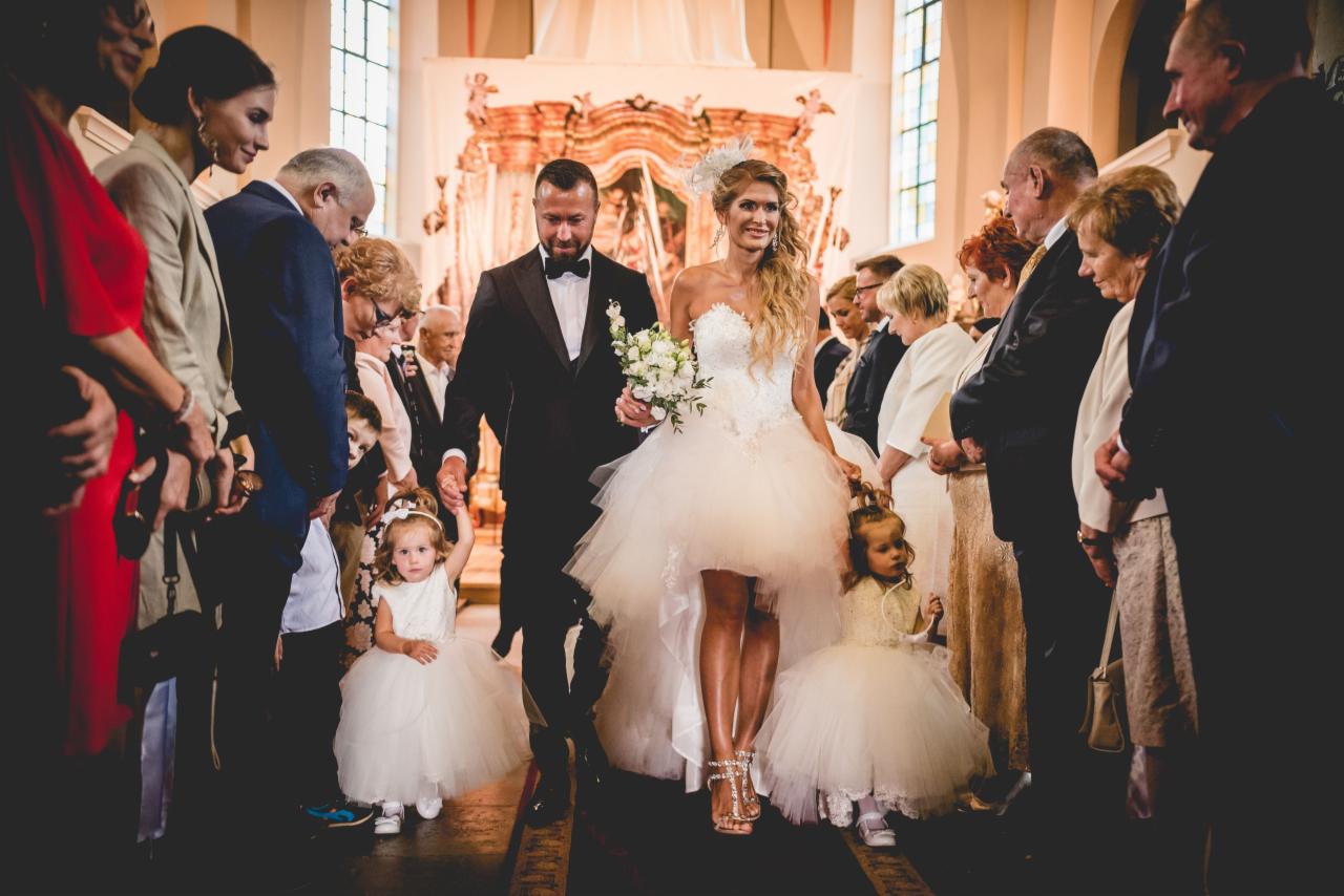 Para młoda w kościele, prowadzona przez dwie małe dziewczynki w białych sukienkach, wśród zgromadzonych gości.