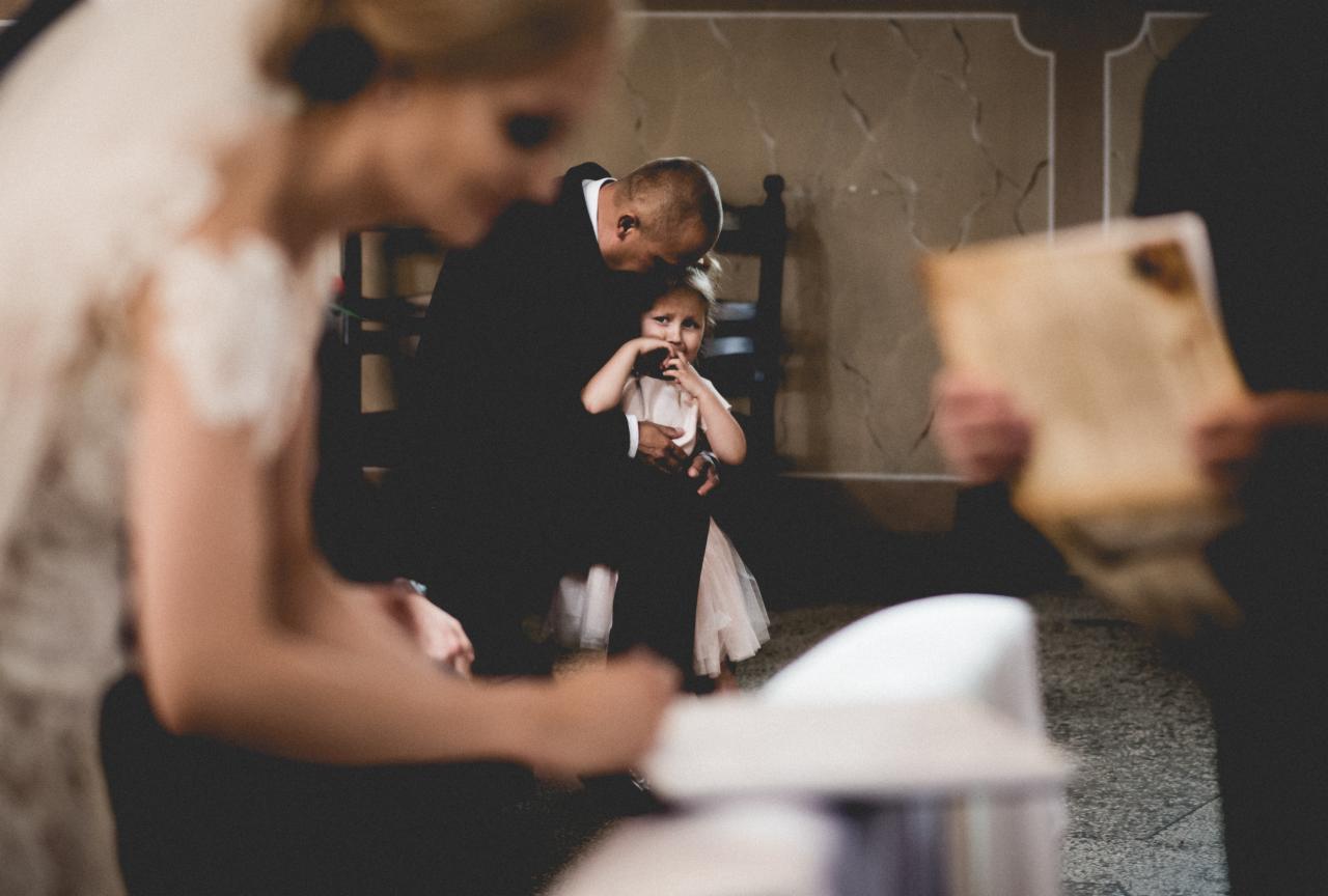 Wzruszony ojciec w czarnym garniturze przytula zapłakaną dziewczynkę w sukience podczas uroczystości ślubnej w kościele.