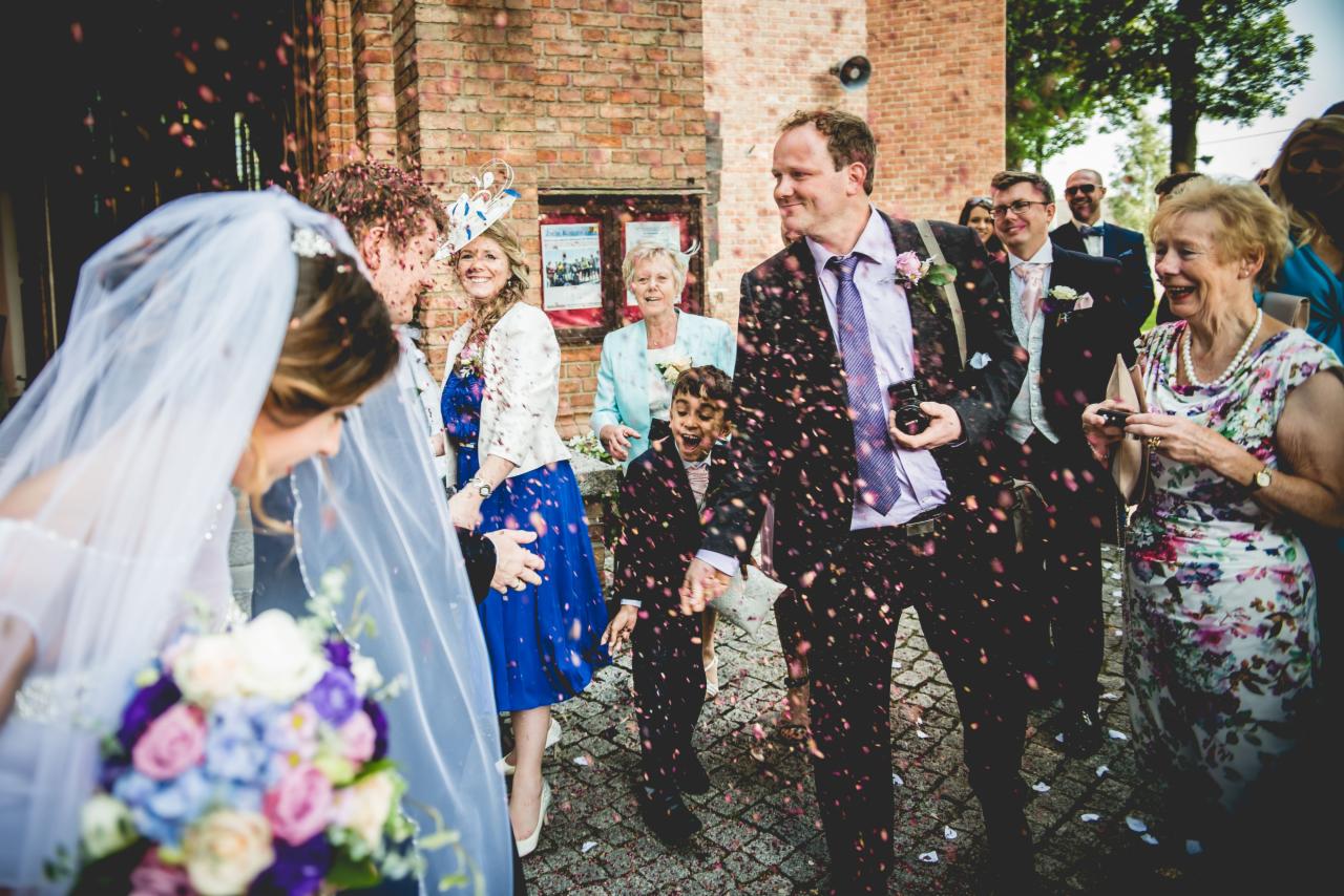Para młoda wychodzi z kościoła obsypana konfetti, goście weselni uśmiechnięci, w tle ceglany budynek.