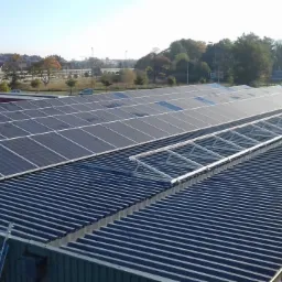 Rząd paneli słonecznych zamontowanych na dachu blaszanym, częściowo z widocznym stelażem montażowym, z drabiną opartą o krawędź dachu i widokiem na zabudowania w tle.