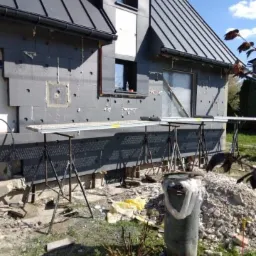 Dom jednorodzinny w trakcie prac związanych z izolacją zewnętrzną, widoczne płyty styropianowe na ścianach, rusztowania, oraz bałagan na posesji.