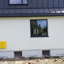 Jasna elewacja domu z ciemną stolarką okienną i dachem, widoczna żółta skrzynka gazowa i małe otwory wentylacyjne przy gruncie.