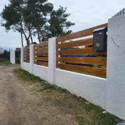 Nowoczesne ogrodzenie posesji z drewnianymi przęsłami i białymi, tynkowanymi słupkami, z czarną skrzynką na listy z motywem trąbki.