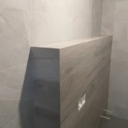 Obudowa stelaża WC z płytek imitujących drewno, dopasowanych do szarych płytek ściennych w łazience, widoczna puszka instalacyjna i fragment instalacji wodnej.