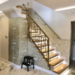 Nowoczesne schody drewniane z podświetleniem LED, metalową balustradą o geometrycznym wzorze, ściana pokryta tapetą w morskim stylu, marmurowe płytki podłogowe i czarny taboret.