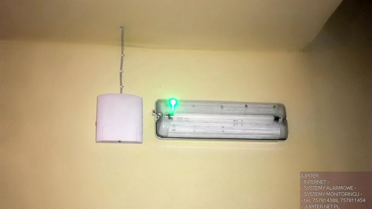 Lampa sufitowa i awaryjne oświetlenie LED z zieloną kontrolką na kremowej ścianie, widoczne logo firmy JUPITER z usługami internetowymi, systemami alarmowymi i monitoringiem.