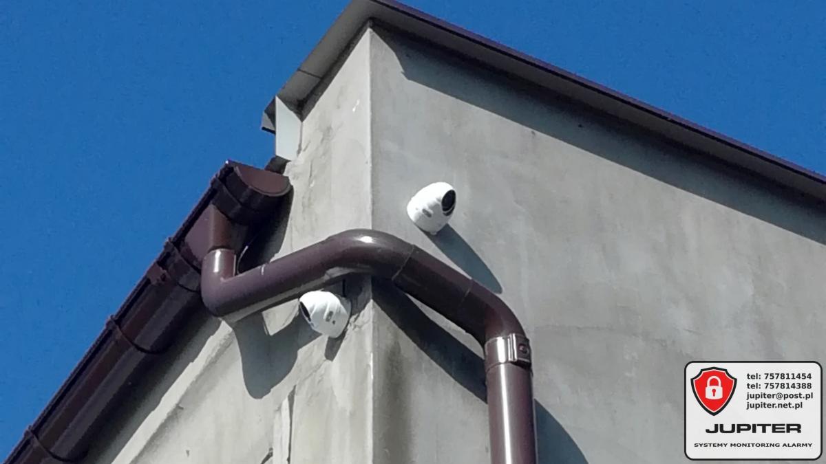 Dwie białe kamery monitoringu zamontowane na rogu budynku z szarą elewacją, obok brązowej rynny, na tle błękitnego nieba. Widoczne logo firmy Jupiter.