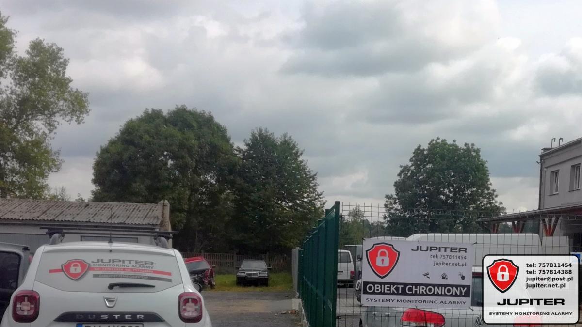 Teren firmy ochroniarskiej Jupiter z zaparkowanymi samochodami, w tym białym Dusterem z naklejką reklamową firmy na tylnej szybie oraz tablicą informacyjną 'Obiekt Chroniony' na ogrodzeniu.