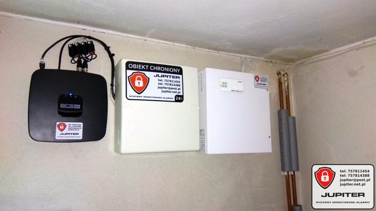 Zestaw urządzeń zabezpieczających: centrala alarmowa BCS, moduł zasilania awaryjnego i skrzynka z napisem 'Obiekt Chroniony Jupiter' na ścianie, widoczne kable i rury instalacyjne.