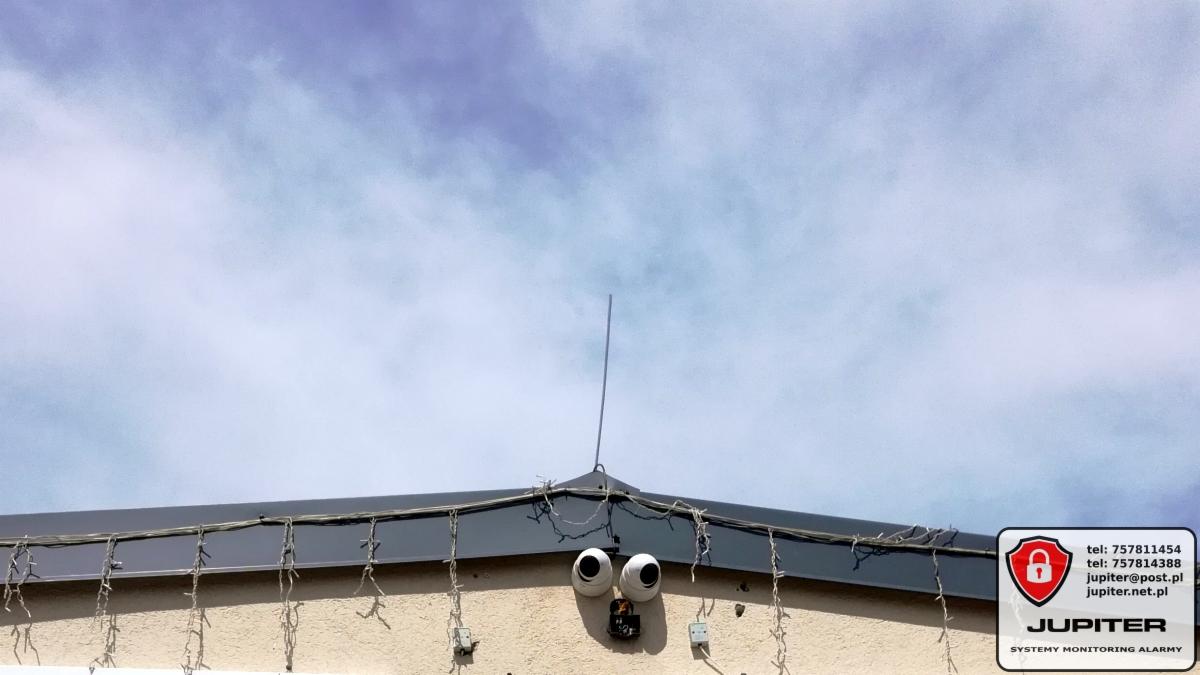 Dwie kamery monitoringu zamontowane na elewacji budynku pod dachem, z widoczną anteną i logo firmy Jupiter w rogu.