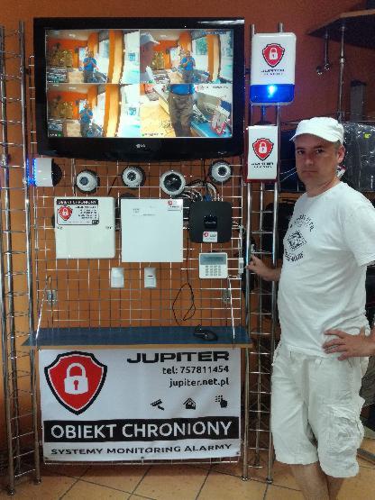 Prezentacja systemów zabezpieczeń na metalowym stojaku: monitor z podglądem z kamer, czujniki, centralka alarmowa, sygnalizatory świetlne, baner z logo Jupiter i hasłem 'Obiekt Chroniony', obok...